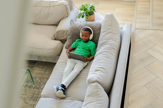 African American boy reclining on gray sofa in living room wearing white headphones holding tablet