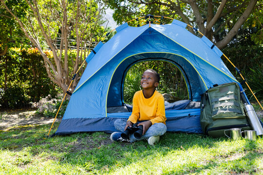 African American boy sitting at tent in yellow shirt holding binoculars with backpack and thermos