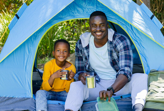 African American father and son sitting at blue tent opening holding mugs, touching food container