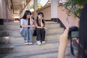 Two teenage girls posing for a photo on school stairs while a friend takes their picture, showing friendship, youth lifestyle, and candid moments in a campus setting.