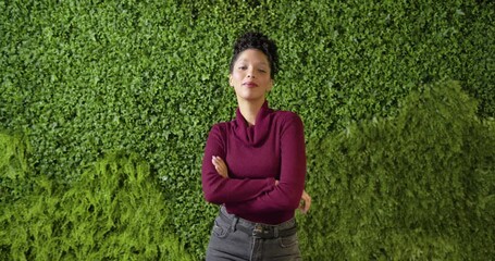 Woman in twenties greeting camera, unfolding arms and laughing before plant wall to engage viewer - Powered by Adobe