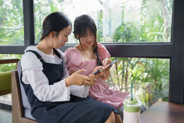 Fototapeta premium Two young women sitting indoors using smartphones together. Concept of social media, digital lifestyle, friendship, connection, technology, and modern communication.