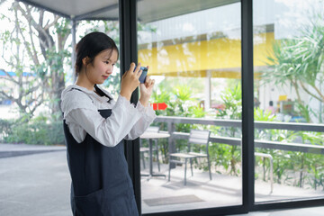 Fototapeta premium Teenage girl standing and taking a photo with her smartphone inside a café, expressing creativity, focus, and modern digital lifestyle in a bright natural environment