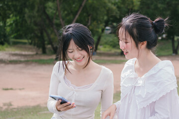 Obraz premium Two teenage girls smiling while looking at a smartphone outdoors, enjoying a bright and relaxed moment together in a natural green environment.