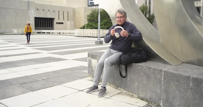 Man arriving, sitting by sculpture in plaza, getting headphones, phone, woman passing, copy space