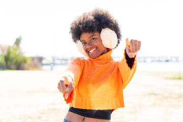 African American girl wearing winter muffs at outdoors points finger at you while smiling