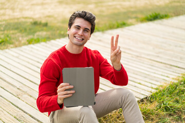 Fototapeta premium Young handsome man holding a tablet at outdoors smiling and showing victory sign