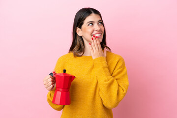 Young Italian woman holding a coffee maker isolated on pink background looking up while smiling