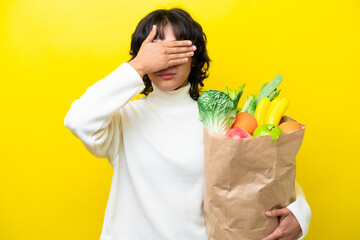Young Argentinian woman holding a grocery shopping bag isolated on yellow background covering eyes by hands. Do not want to see something