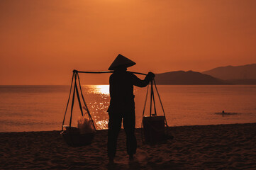 Silhouette of a woman in a traditional Vietnamese hat carrying fruits along the seashore at dawn.