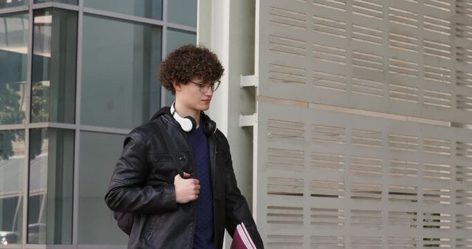 Entering frame, man walking along sidewalk, wearing headphones and carrying backpack and red folder