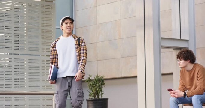 Asian male student spotting peer, walking with books, strapping backpack, scanning facade on campus