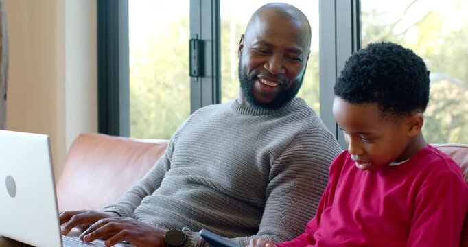 African American father and son showing tablet on sofa at home glancing smiling returning to laptop