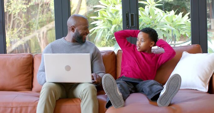African American father and son pausing on laptop after son gesturing question, laughing on couch