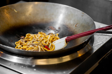 A close-up shot of a white and red spatula mixing delicious stir-fried noodles with bean sprouts and chili in a large metal wok over a hot stove.