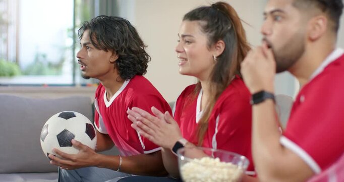 Diverse friends wearing red jerseys on sofa at home holding ball and popcorn, celebrating goal