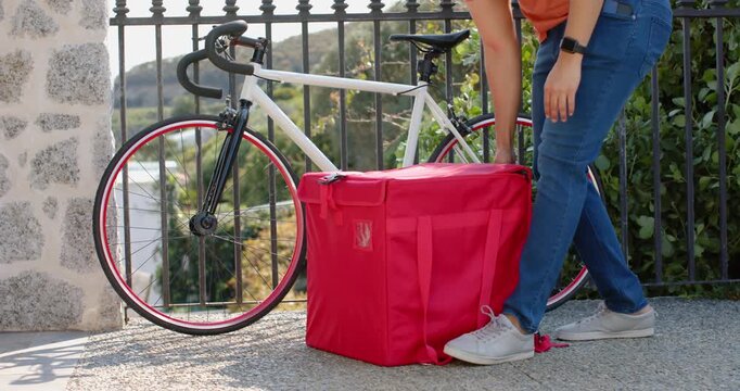 courier approaching bicycle and bag on walkway, lifting bag onto back and pulling bike for delivery