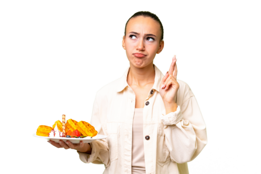 Young Arab woman holding waffles over isolated background with fingers crossing and wishing the best