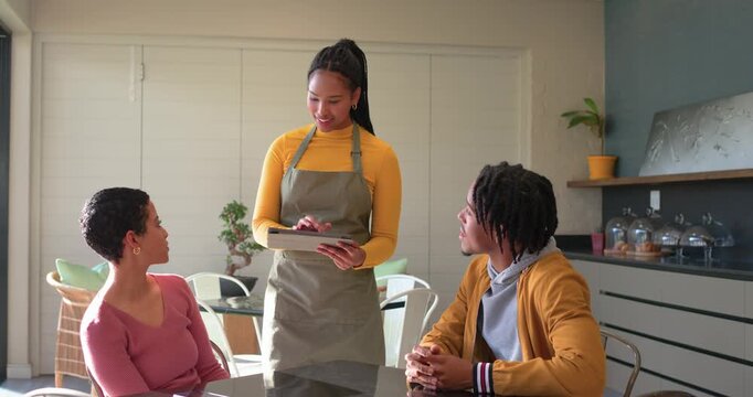 Woman server in apron approaching cafe table greeting couple, presenting menu tablet to take order