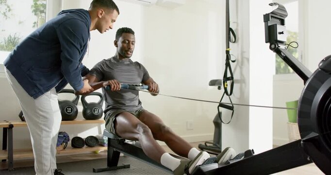 Trainer guiding mid adult African American man rowing on rower for posture correction in home gym