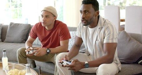 Diverse male friends picking controllers on home sofa as game loading, strategizing to win, smiling