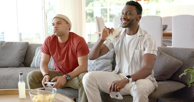 Diverse male friends settling on sofa at home, grabbing gamepads, sipping drinks and competing