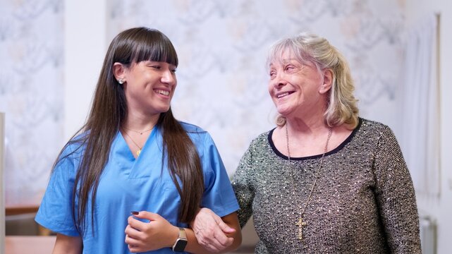 Nurse assisting elderly woman walking, smiling in a nursing home