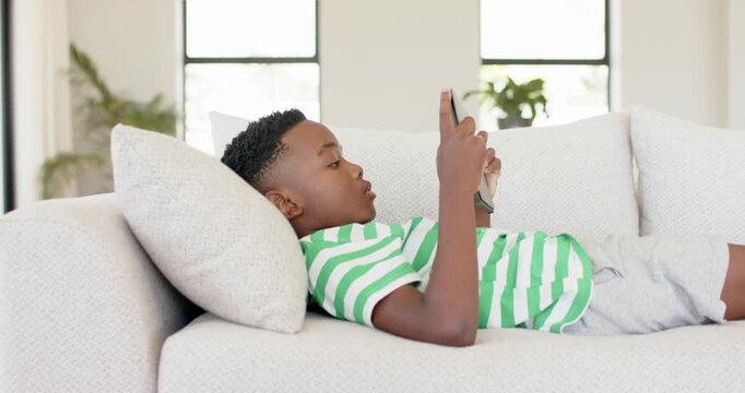 African American boy lifting handheld device from sofa cushions in living room scrolling content
