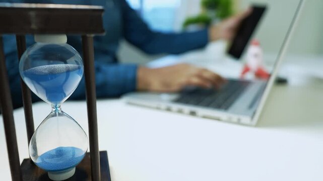Close-up of an hourglass with blue sand on a desk, person using a tablet in the background
