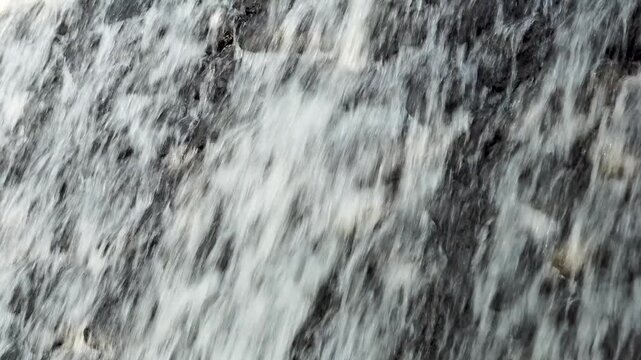 A close-up of a curtain of waterfall cascading down a dark, rough rock face. Focus on the rapid vertical movement of the white water, creating a dynamic and repetitive visual texture.
