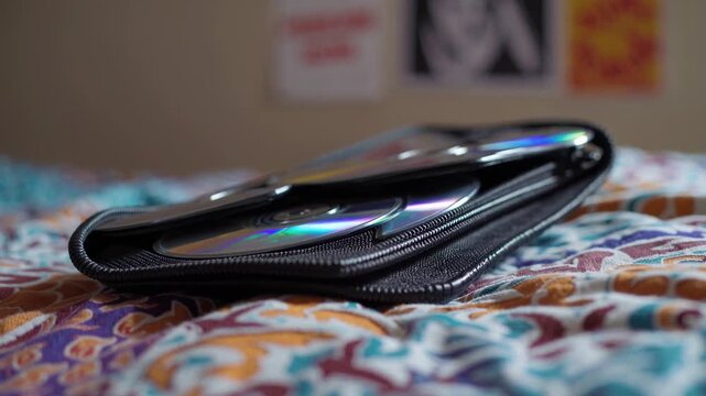 Portable black cd wallet holding multiple iridescent compact discs with colorful reflections, resting on a patterned bedspread, evoking nostalgia for vintage music and data storage