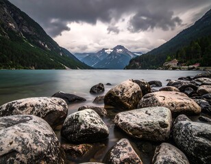Serene mountain lake scene with boulders in foreground, dramatic clouds, and forested slopes