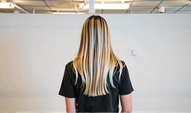 Woman with long blonde and dark striped hair seen from behind standing against a white brick wall