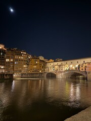 Obraz premium The Ponte Vecchio glows under the night sky, its historic arches reflecting in the Arno and carrying centuries of Florentine charm.