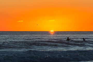 Vivid ocean sunset with the sun touching the horizon, illuminating the sky in intense orange tones while two swimmers appear as silhouettes amid the gentle waves. Caravelos beach, Portugal