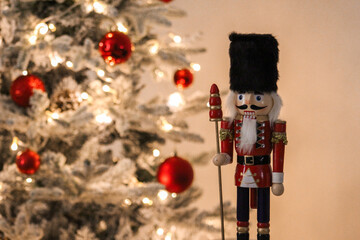 Wooden nutcracker toy standing next to a decorated Christmas tree with warm lights and red ornaments.