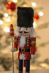 Vertical image of a colorful nutcracker figure placed near a glowing Christmas tree with red ornaments.
