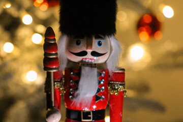 Wooden nutcracker guard with bright colors and fluffy hat photographed in front of glowing Christmas lights.