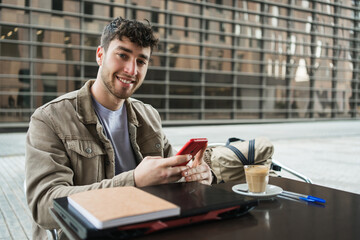 Young man smiling at outdoor cafe while holding smartphone and laptop