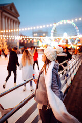 Beautiful tourist in winter clothes with warm drink in hand stands near skating rink at Christmas...
