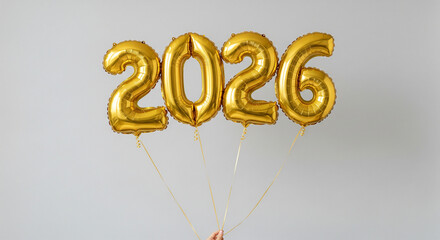 A hand holding golden metallic helium balloons shaped as the numbers "2026" against a plain light gray background. The shiny gold balloons are tied with golden ribbons, creating a festive New Year.