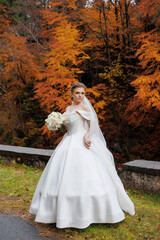 Fototapeta premium Bride in elegant white dress stands near autumn trees holding bouquet during outdoor wedding