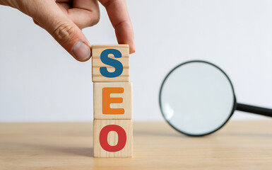 Hand carefully stacks wooden blocks spelling SEO next to a magnifying glass symbolizing search engine optimization strategy