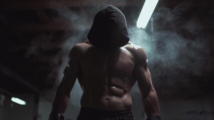Muscular boxer with hood up standing in a dark gym surrounded by smoke. Sweaty male fighter preparing for combat. Intense fitness and strength concept
