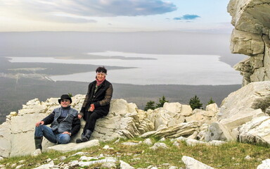 Mature tourists, a man and a woman, sit on the rock of the Zyuratkul ridge in the background of the mountain lake Zyuratkul