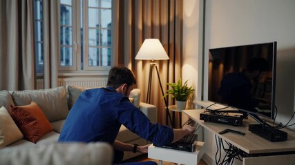 Professional technician in overalls connecting multiple cables to a set top box, troubleshooting electronics entertainment system in a comfortable home living room