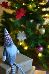 Cozy Christmas scene with a small Nordic gnome sitting on wrapped gifts under a decorated tree. Kraft paper boxes, soft handmade toy and natural holiday decor create a warm festive atmosphere at home.