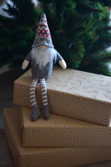 Cozy Christmas scene with a small Nordic gnome sitting on wrapped gifts under a decorated tree. Kraft paper boxes, soft handmade toy and natural holiday decor create a warm festive atmosphere at home.