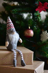 Cozy Christmas scene with a small Nordic gnome sitting on wrapped gifts under a decorated tree. Kraft paper boxes, soft handmade toy and natural holiday decor create a warm festive atmosphere at home.