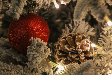 Festive close-up of a red glitter ornament and a pine cone on a snow-covered Christmas tree with warm holiday lights.
